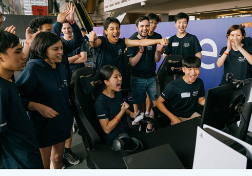 A group of young people standing around a screen and celebrating as a young woman wins a videogame competition. 