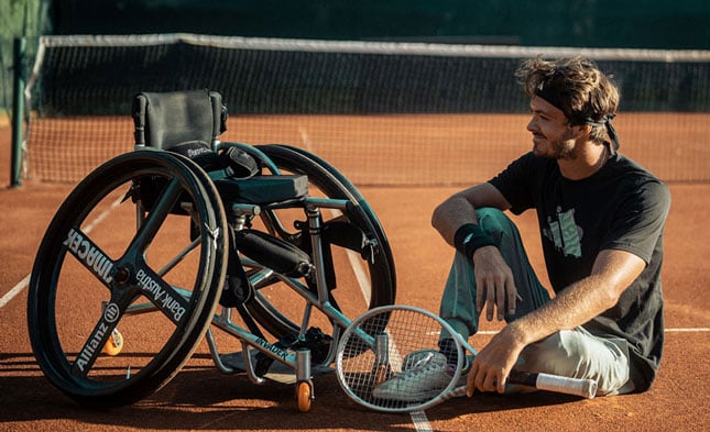 Nico Langmann sitting on tennis court beside a specialized sports wheelchair.