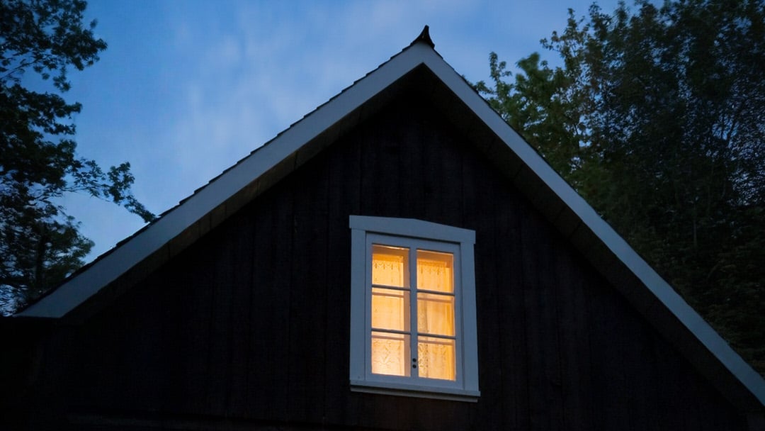 Dunkles Holzhaus mit beleuchtetem Fenster in der Dämmerung, umgeben von Bäumen.