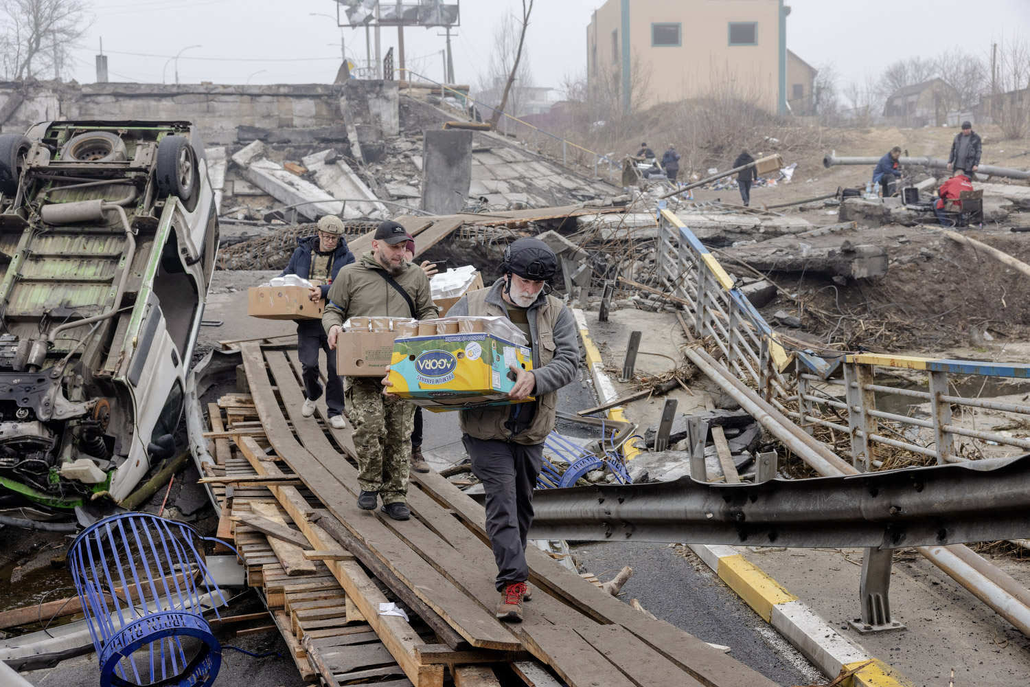 Drei Mitarbeiter von World Central Kitchen gehen über Holzplanken im total zerstörten Bucha und tragen Kisten mit Essenvorräten