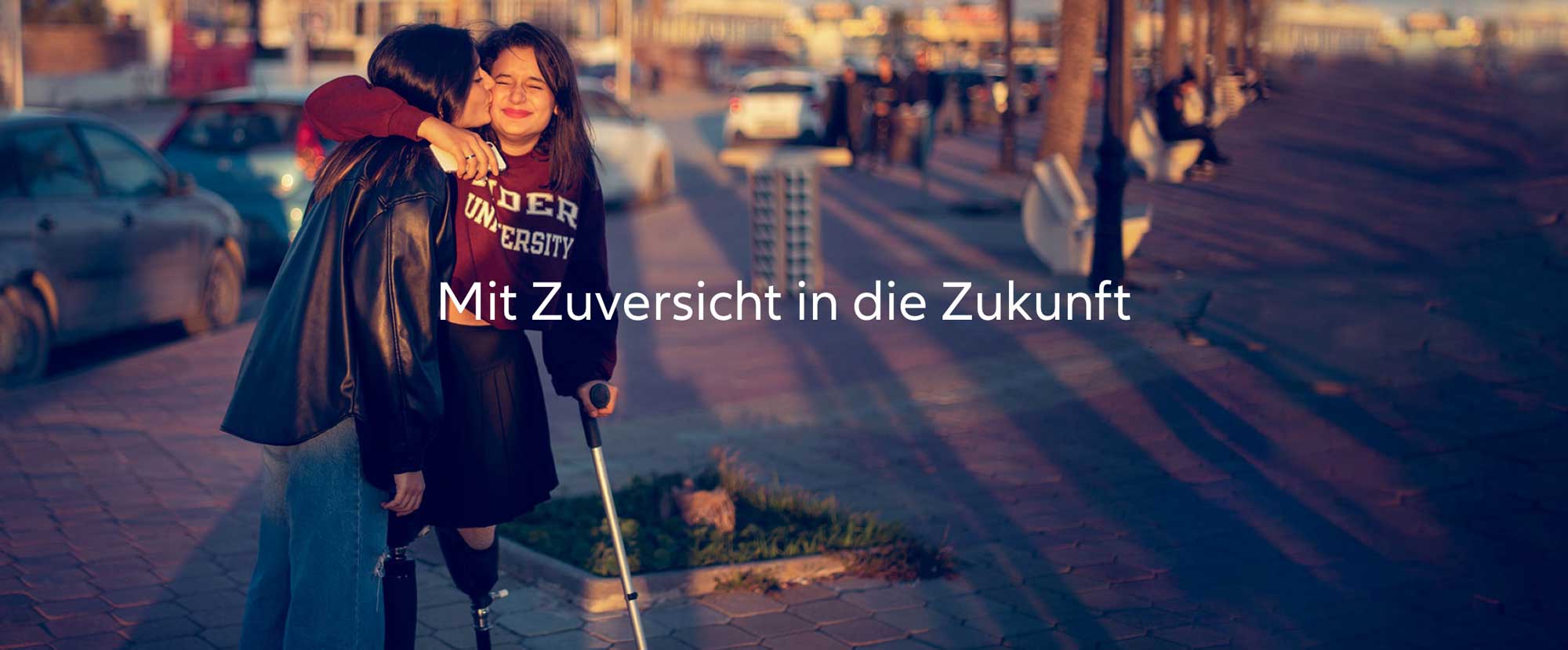 Zwei Freunde, eine davon mit Beinprothese, umarmen sich, öffentlicher Ort, Abendsonne – Mit Zuversicht in die Zukunft