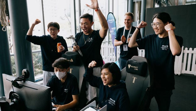 A group young people standing around a screen and celebrating the result of a videogame competition.
