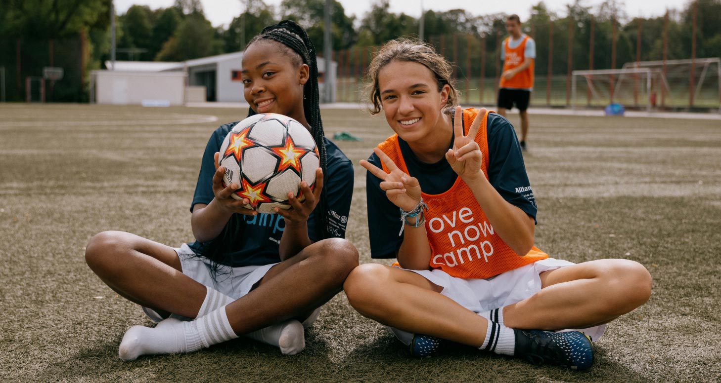 Zwei Teilnehmerinnen auf dem Fußballrasen sitzend, einen Fußball haltend, lachen fröhlich in die Kamera