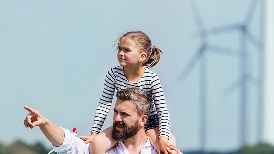 Vater mit Tochter auf der Schulter Finger in Richtung Zukunft zeigend, im Hintergrund Windräder