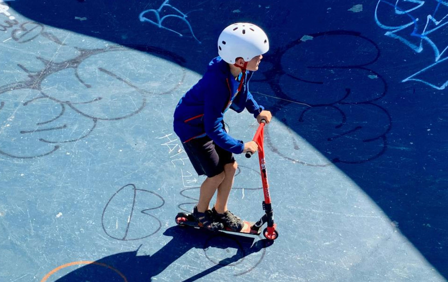 Little boy, wearing a helmet, rides his scooter on a skater training ground