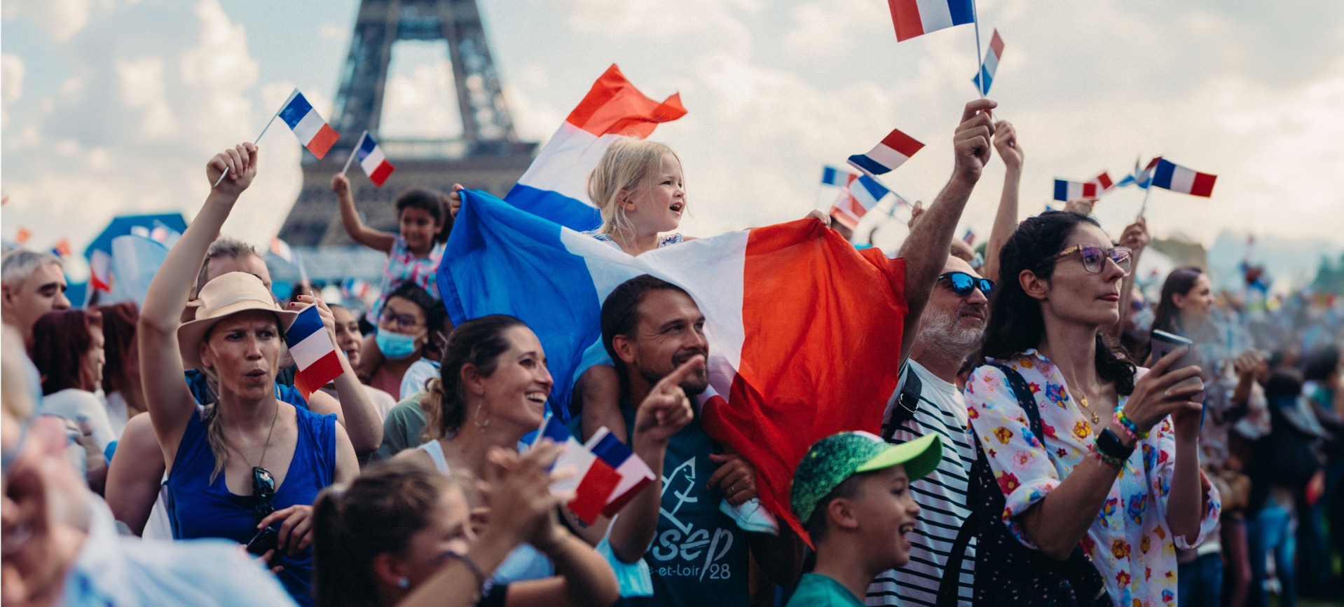 Crowd of cheering fans, girl on her father's shoulders with many waving France flags, the Eiffel Tower in the background
