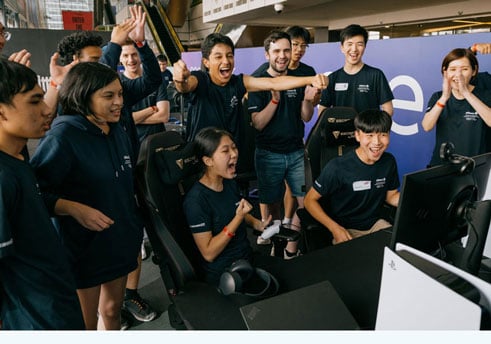 A group of young people standing around a screen and celebrating as a young woman wins a videogame competition. 