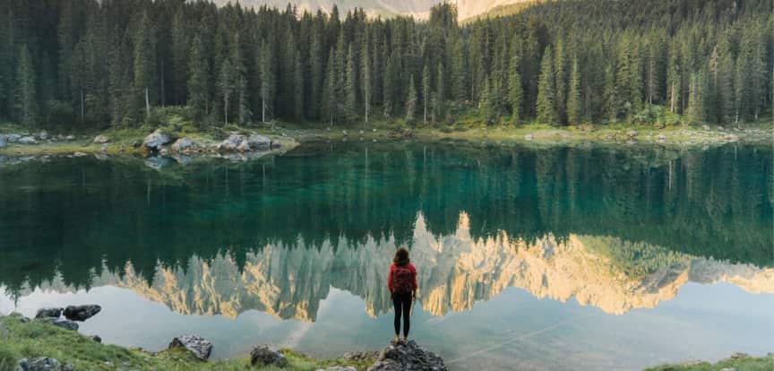 A woman stands in front of a scenic lake with mountains reflected on its surface 