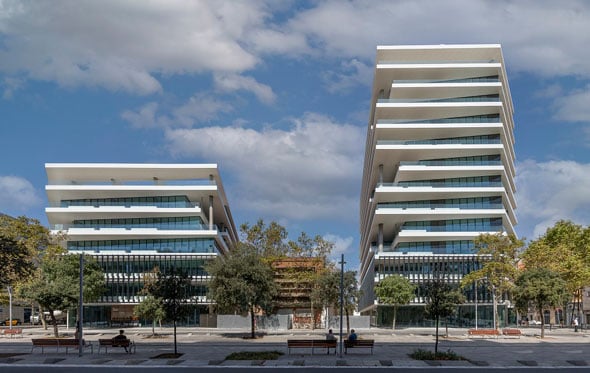 Two buildings built in the same style. Gleaming white floors with large open balconies and bluish windows give a very clean, modern impression. The building on the left has 7 stories; the one on the right has 13 stories. Blue sky with some scattered white clouds serves as serene backdrop.