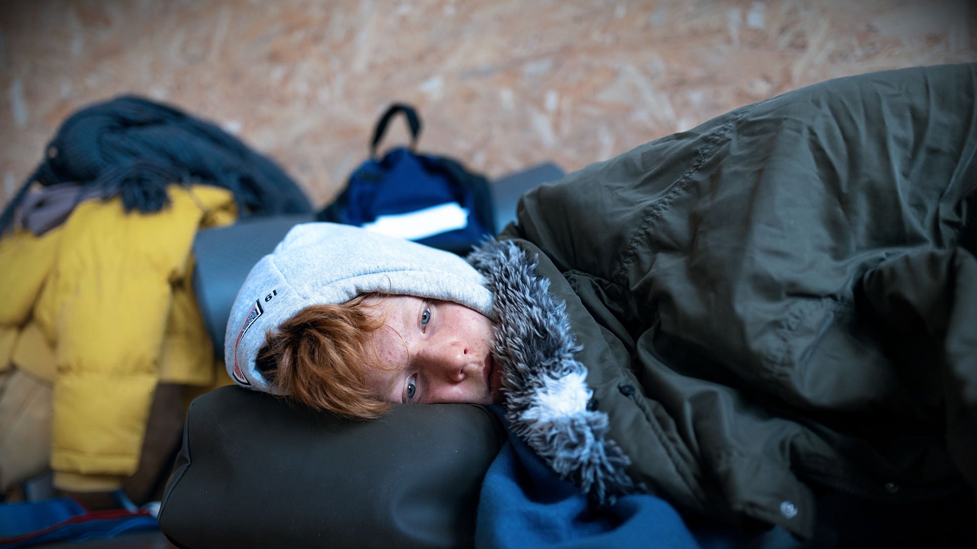 Child with red hair lying in a sleeping bag, surrounded by coats and bags.