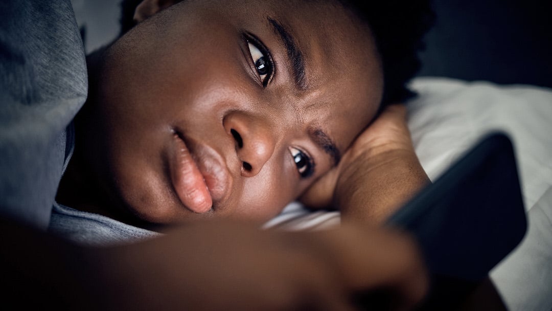 Person lying in bed at night, looking thoughtfully at a smartphone.