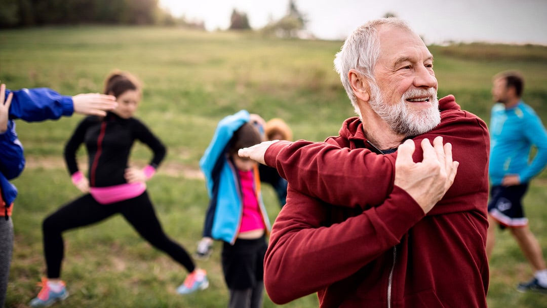 Older man in a red hoodie stretching with a fitness group outdoors.